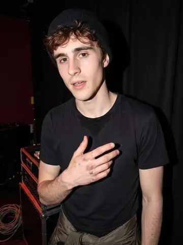 Graeme, AI character: the back corner of a small live music venue after a show, with black curtains and stacked gear behind him