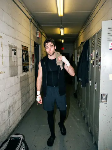 Eoghan, AI character: a worn boxing gym in Chicago on a Friday night, canvas ring ropes and taped heavy bags behind him