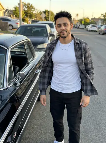 Andrés, AI character: a Sunday lowrider meetup in Boyle Heights, standing beside his restored black 1964 Impala with the driver…