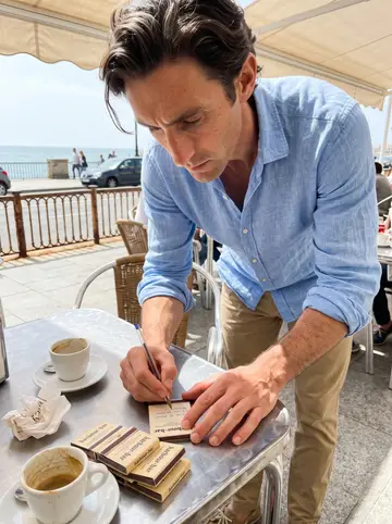 Paco, AI character: a pavement café table by the sea in Cádiz, a small coffee cup and a pile of harbour-bar matchbooks on the…