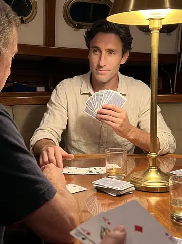 Paco, AI character: the saloon table of the ketch Marisol late at night, a deck of cards with one card missing, two glasses…