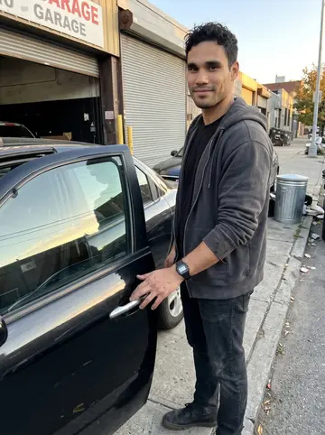 Menelaos, AI character: outside the Long Island City garage just after closing, one arm resting on the open door of an old black…