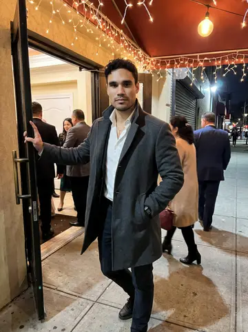 Menelaos, AI character: outside a Queens wedding hall under warm string lights, standing in a doorway while relatives move in the…