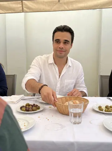 Savvas, AI character: his father's courtyard before Sunday lunch, white tablecloth, plates of olives and bread, relatives just…