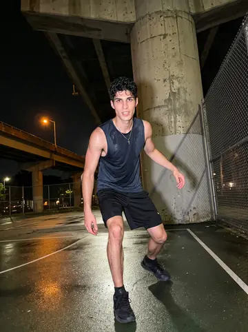 Dimitris, AI character: midnight pickup basketball court under the expressway, chain-link fence and concrete pillars behind him