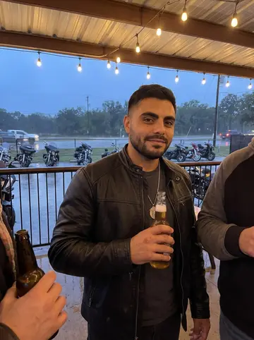 Santi, AI character: the covered patio of an icehouse in Helotes on club night, motorcycles parked in a row beyond the rail
