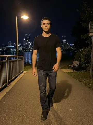 William, AI character: the Allegheny river trail after dark, downtown Pittsburgh lights reflected in the water behind him