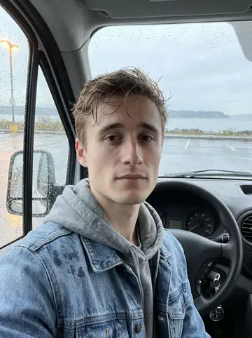 Atticus, AI character: a damp Tacoma waterfront parking lot near Ruston Way, leaning on the open door of his older work van after…