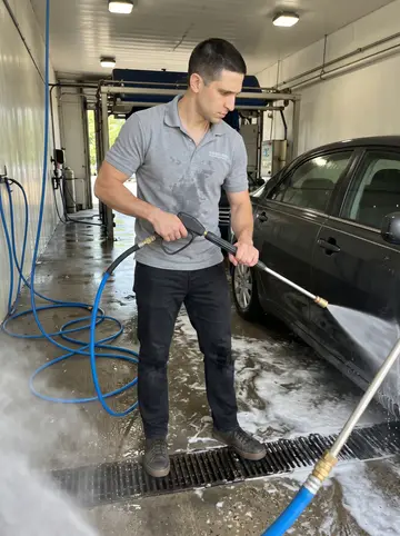 Predrag, AI character: inside the car wash tunnel during a slow afternoon shift, blue hoses and wet floor around him