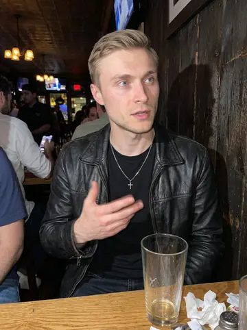 Dmitry, AI character: a crowded neighborhood bar near Brighton Beach after service, dark wood wall, half-empty pint glasses, and…