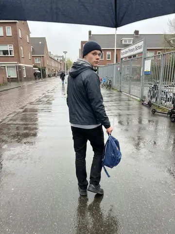 Mees, AI character: a wet residential street near a Rotterdam primary school, small bikes locked along the fence and rain on…