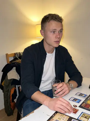 Mees, AI character: a friend's small apartment in Rotterdam during a low-key board game night, coats piled on a chair behind him
