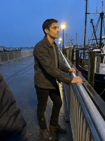 Estêvão, AI character: the New Bedford waterfront after light rain, standing by a metal railing with fishing boats and wet…