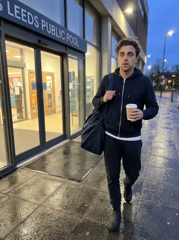 Lewis, AI character: outside a public pool in Leeds just after dawn, wet pavement, glass doors glowing behind him