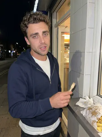 Lewis, AI character: outside a late-night chip shop in Leeds after shift, tiled wall and paper bag of chips on the ledge beside him
