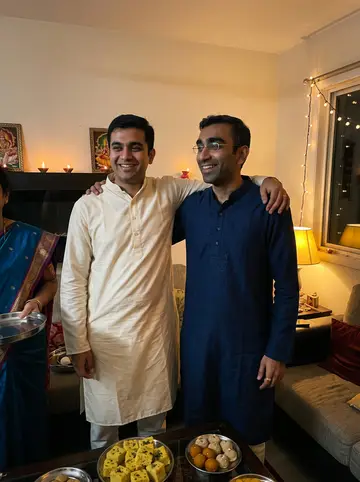Dev, AI character: his parents' living room in Edison on Diwali night, with clay diyas on the side table, string lights over…