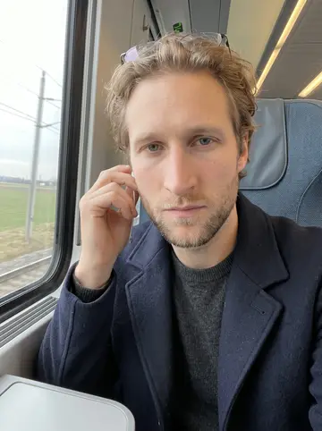 Felix, AI character: concrete steps on the Donauinsel shore, the grey-green Danube behind him, bare trees and a distant bridge