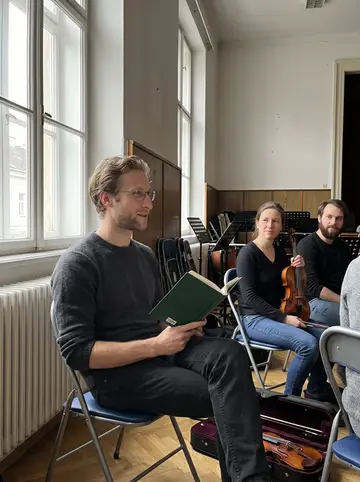 Felix, AI character: Wednesday rehearsal of an amateur orchestra in a school hall in Ottakring, music stands, folding chairs, a…