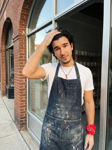 Gianni, AI character: in the open doorway of his small pottery workshop in Bologna, a former bike shop with a glass front and…