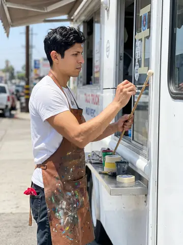 Raúl, AI character: beside a taco truck in Boyle Heights while he hand-letters the service window trim