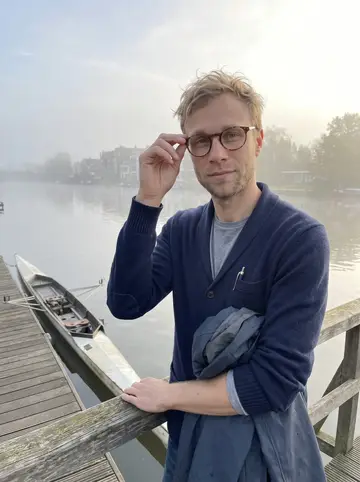 Jonah, AI character: the end of a boathouse dock on the Willamette in Portland, leaning on the rail beside his racked single scull