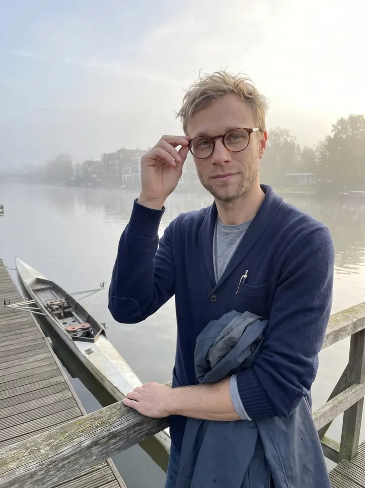 Jonah, AI character: the end of a boathouse dock on the Willamette in Portland, leaning on the rail beside his racked single scull