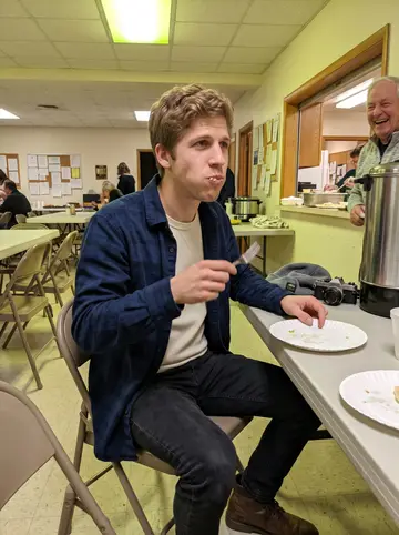 Mark, AI character: pierogi night in a Polish church basement, folding tables, paper plates, and a coffee urn behind him