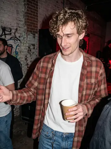 Beau, AI character: a small warehouse punk show in East Austin, painted brick wall and a few friends blurred behind him