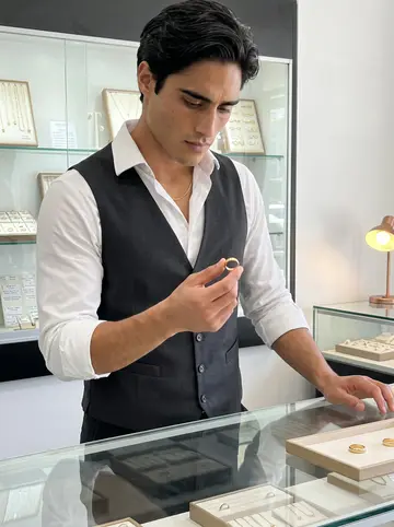 Alexis, AI character: inside the quiet jewelry shop before opening, seated at a glass counter with trays of rings and a small…