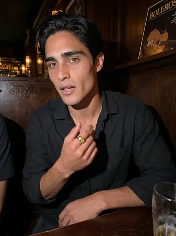 Alexis, AI character: a small low-key bar with old boleros playing, crowded table edge and dark wood wall behind him
