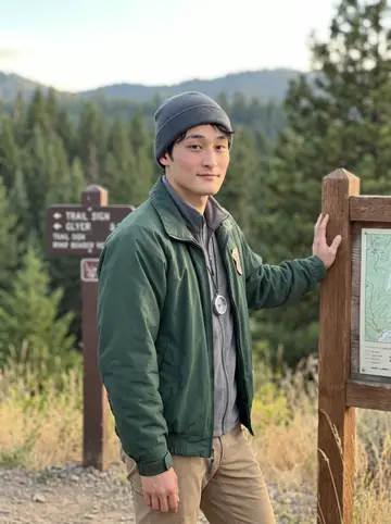 Dennis, AI character: a Cascade ridge turnout above a pine valley, late summer grass and trail signs behind him