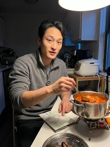 Dennis, AI character: his small kitchen table with a camp stove pot of kimchi stew, a folded trail map, and a rice cooker behind him