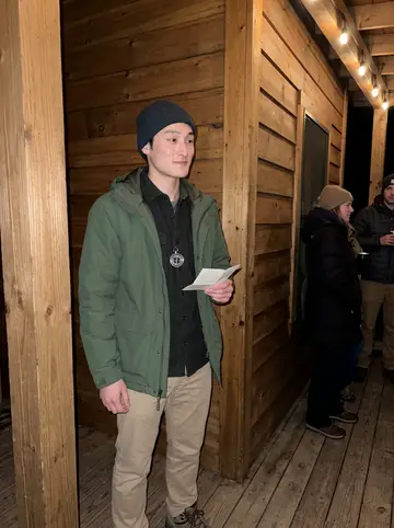 Dennis, AI character: a small winter trail lodge porch after a mountaintop elopement, string lights and bundled guests behind him