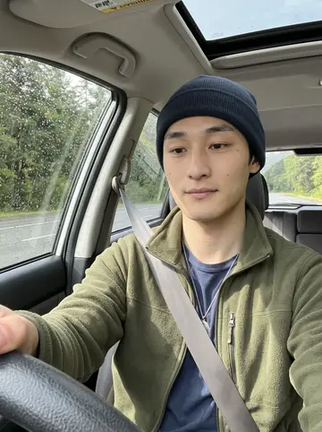Dennis, AI character: inside his parked old Subaru at a scenic pullout after rain, wet evergreens and a cloudy highway visible…