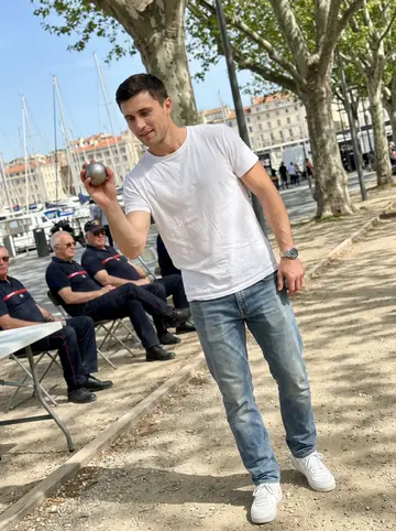 Idriss, AI character: a pétanque court near the Vieux-Port with retired firefighters sitting on folding chairs in the background
