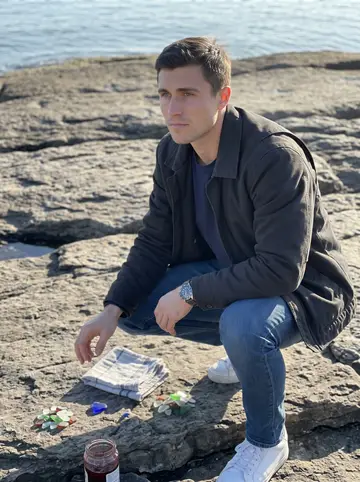 Idriss, AI character: flat rocks near the Vieux-Port, cold clear water behind him, a few glass pieces spread on a folded cloth