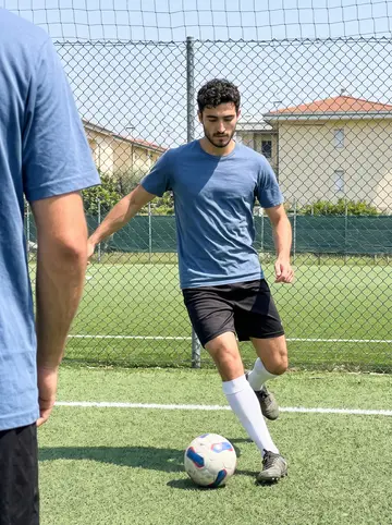Ottavio, AI character: small outdoor five-a-side football pitch on the edge of Bologna, chain-link fence and low apartment blocks…