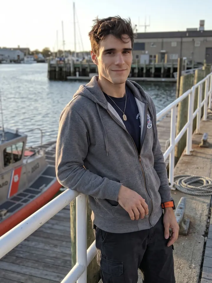 Giosuè, AI character: outside the Gloucester Coast Guard station near the pier, leaning against a white railing with rescue…