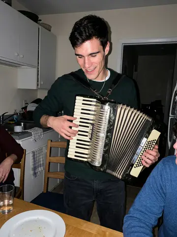 Giosuè, AI character: a crowded Gloucester apartment kitchen after dinner, cousins around the table while he tries to play his…