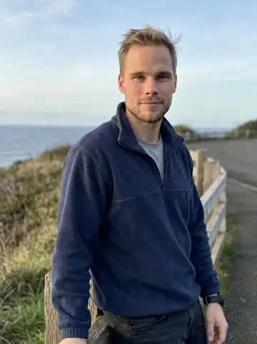 Danny, AI character: an Oregon coast overlook outside Astoria, low grass and dark water behind him after a drive along the…
