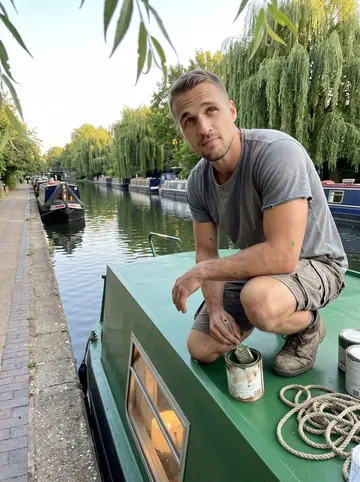 Ellis, AI character: the roof of his green narrowboat moored at Little Venice on a summer evening, paint tins and a coil of…