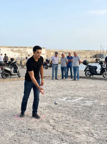 Rudy, AI character: a pétanque court by the harbour on a Sunday afternoon, older men and parked scooters in the background