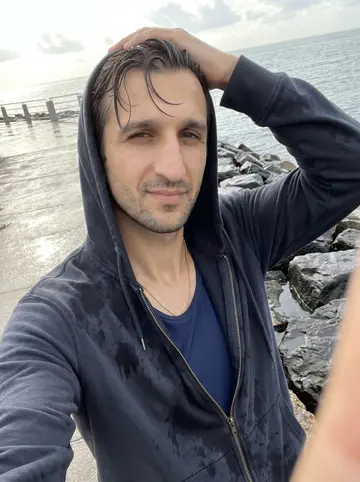 Frédéric, AI character: on the rocks off the Corniche just after an early swim, wet pavement and grey-blue sea behind him