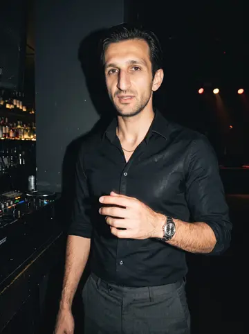Frédéric, AI character: inside his club on the Old Port during a small Marseille chanson set, dark bar wall and stage lights…