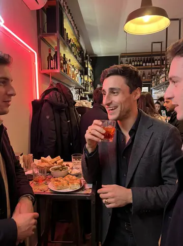 Domenico, AI character: a crowded aperitivo bar on the Navigli canal in Milan, a small high table with glasses of red wine