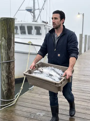 Enzo, AI character: the wooden fish dock at Point Judith at dawn, a trawler tied up behind him, gulls on the pilings