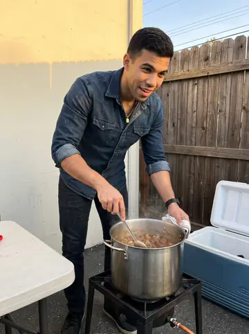 Aarón, AI character: behind the detailing shop on a Friday evening, cooking carne guisada for the crew beside a folding table…