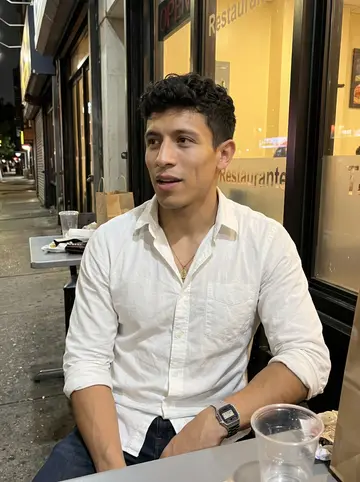 Wilmer, AI character: outside a Colombian restaurant in Jackson Heights after dropping off fruit, sitting at a sidewalk table…