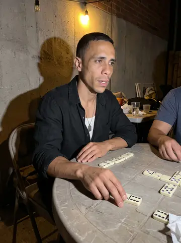 Reinier, AI character: his tío's back patio in Hialeah during Sunday dominoes, plastic table, domino tiles, folding chairs, and…