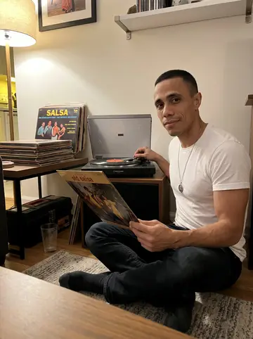 Reinier, AI character: his small apartment living room with salsa records stacked by an old turntable and a toolbox under the…